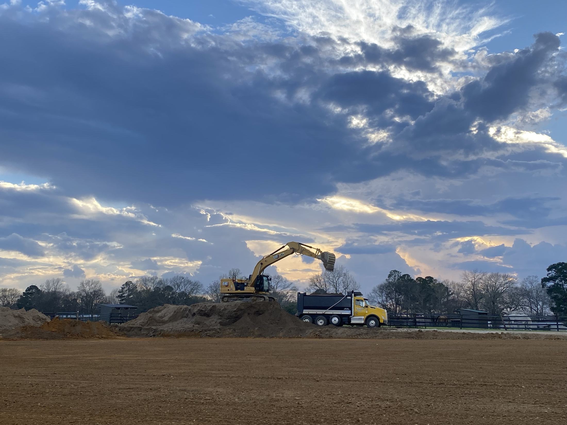 Excavator loading a dump truck on an open site under a dramatic sunset sky