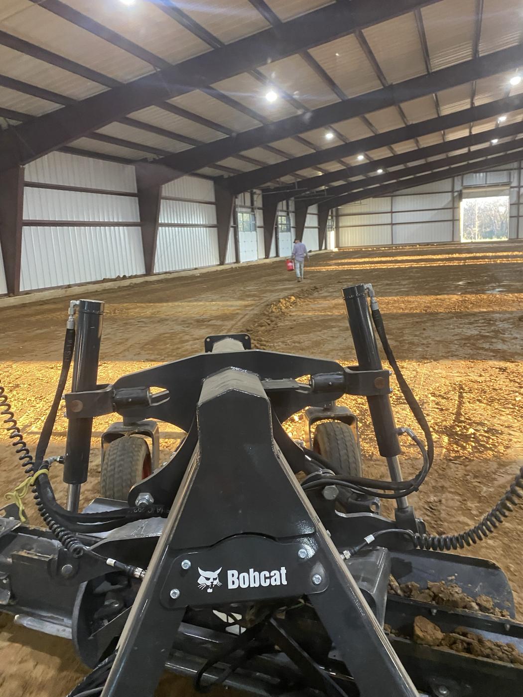 View from the cab of a Bobcat track loader with a laser-grading attachment, inside an arena building