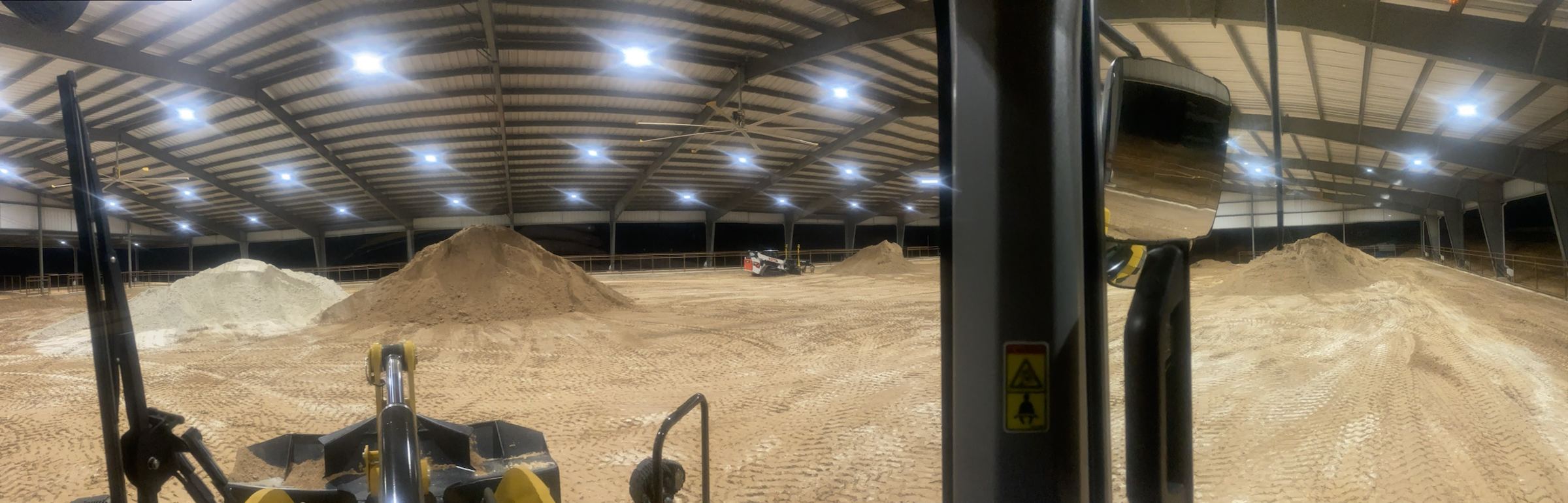 Panorama from the loader cab inside a covered arena, with piles of base and footing material ready to spread