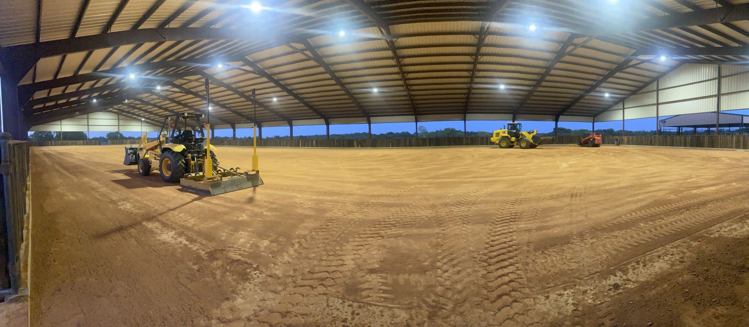 Panorama of a covered arena at dusk: a tractor with a laser box grader setting the base, a CAT loader and track loader beyond